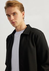 Black zip-up jacket with a flat collar over a white T-shirt. Smooth fabric texture, structured design, and minimal detailing.