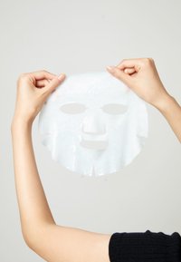 Valkoinen kasvonaamio, jota pidetään käsissä, jossa on leikattuja silmä- ja suunaukkoja, hieman läpikuultava ja kiiltävä pinta, ja se tiputtaa kosteutta.