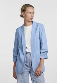 Blazer azzurro chiaro con una silhouette su misura, caratterizzato da un colletto sciallato, maniche lunghe e una texture morbida e tessuta. Abbinato a una camicia bianca.