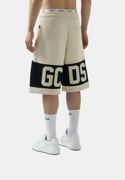 Henkilö, jolla on päällään beigen ja mustan sävyiset shortsit, joissa on takana teksti "GCDS", valkoiset sukat tekstillä ja valkoiset lenkkarit, seisoo selin katsojaan.