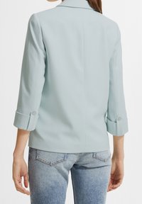 Ljusblå kavaj med en strukturerad design, trekvarts ärmar och knäppta manschetter, kombinerad med ljusblå jeans. Slät tygstruktur.