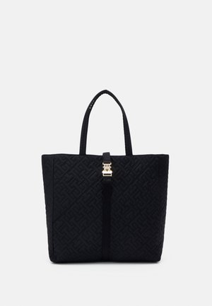 Sac cabas noir matelassé avec motif géométrique embossé, double poignées et fermoir central doré sur une lanière verticale.