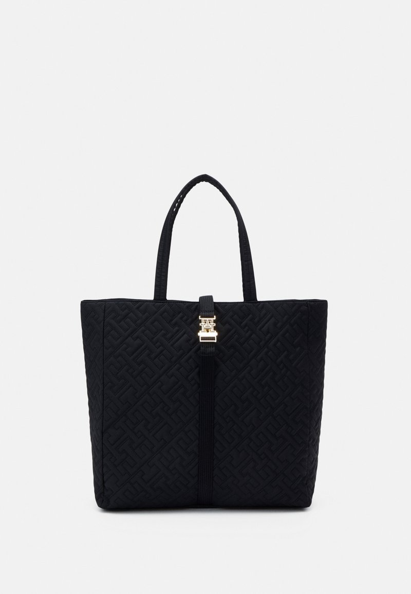 Sac cabas noir matelassé avec motif géométrique embossé, double poignées et fermoir central doré sur une lanière verticale.