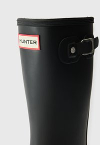 Schwarzer Gummi-Regenschuh mit strukturierter Oberfläche, ausgestattet mit einem rot-weißen Etikett, das mit "HUNTER" gekennzeichnet ist, und einem Seitengurt mit einer silbernen Schnalle.