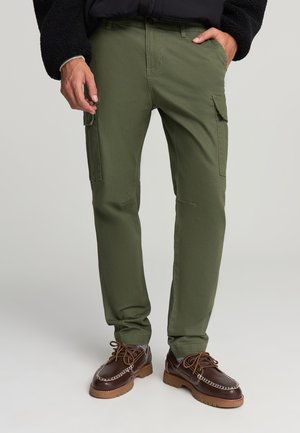 Pantaloni cargo verdi con una vestibilità slim, dotati di due tasche laterali, texture liscia e dettagli di cucitura discreti. Abbinati a scarpe marroni con lacci.