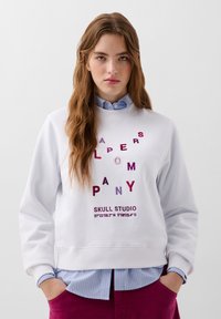 Biała bluza z kolorowymi, haftowanymi literami i współrzędnymi, w połączeniu z niebieską w paski koszulą, noszona z bordowymi spodniami.