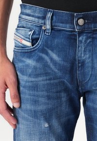 Modré džínsy s vyblednutým umytím, ktoré majú predné vrecko s červenou nášivkou a opotrebovaný detail v oblasti stehna.