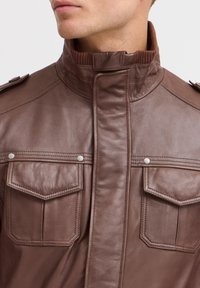 Veste en cuir marron avec un col haut, deux poches poitrine à boutons-pression et épaulettes. Texture lisse avec des détails cousus.