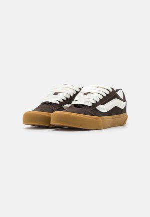 Zapatillas deportivas de ante marrón de caña baja con franjas laterales blancas, que presentan cordones blancos y una suela de goma. Parte superior texturizada y cuello acolchado.