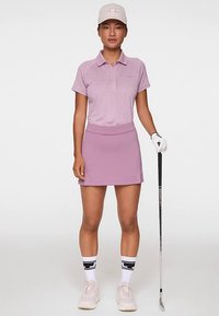 Golfasupistus, jossa on vaaleanpunainen poolopaita ja yhteensopiva hame. Valkoiset sukat mustilla yksityiskohdilla, urheilulliset kengät ja yhteensopiva lippalakki.