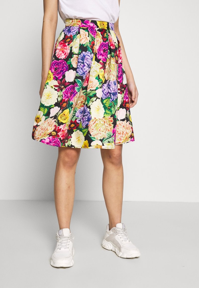 Jupe fleurie longueur genou avec des fleurs multicolores éclatantes, associée à des baskets blanches et un haut blanc rentré, portée par une personne debout.