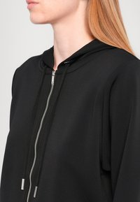Zwarte zip-up hoodie met een trekkoordcapuchon. Glad materiaal, minimalistisch ontwerp, met een zilveren rits en zilveren hardwareaccenten.