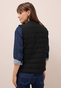 Gilet imbottito nero con design a righe orizzontali, indossato sopra una camicia di jeans blu con le maniche arrotolate. Il tessuto sembra leggero e liscio.