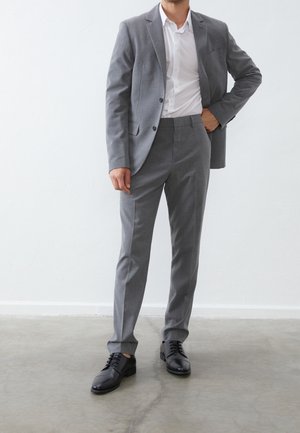 Costume gris composé d'un blazer sur mesure et d'un pantalon assorti, associé à une chemise blanche et des chaussures à lacets noires. Tissu lisse et coupe structurée.
