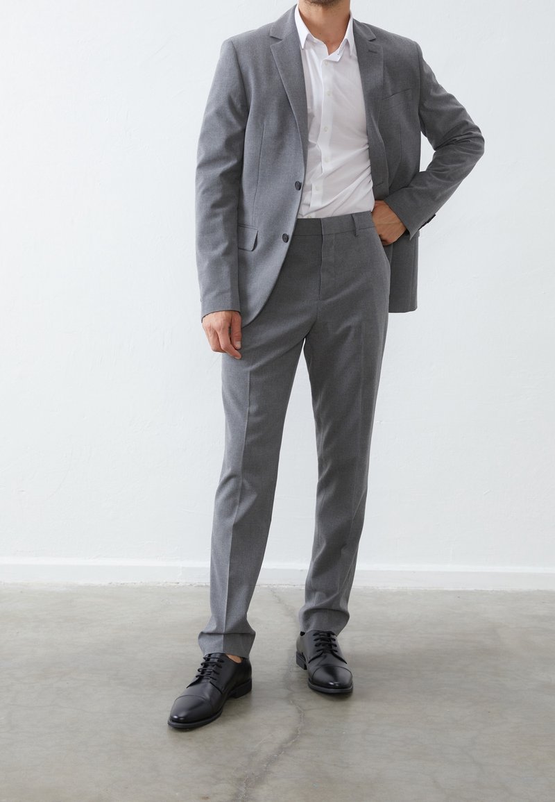 Costume gris composé d'un blazer sur mesure et d'un pantalon assorti, associé à une chemise blanche et des chaussures à lacets noires. Tissu lisse et coupe structurée.