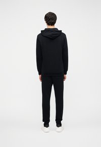 Μαύρο φούτερ με κουκούλα και μπροστινή τσέπη, casual εφαρμογή, ριμπ μανσέτες, κουκούλα με κορδόνι· συνδυασμένο με μαύρο αθλητικό παντελόνι και λευκά αθλητικά παπούτσια.