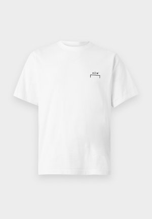 Mees seisab käed taskutes, seljas valge lühikeste varrukatega särk, helehallad püksid ja mustad jooksujalatsid valgel taustal.