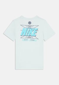 Svijetloplava majica s kratkim rukavima s velikim "NIKE" logom u podebljanoj cijan boji, s geometrijskim uzorcima i natpisom "EST 1908" na leđima.