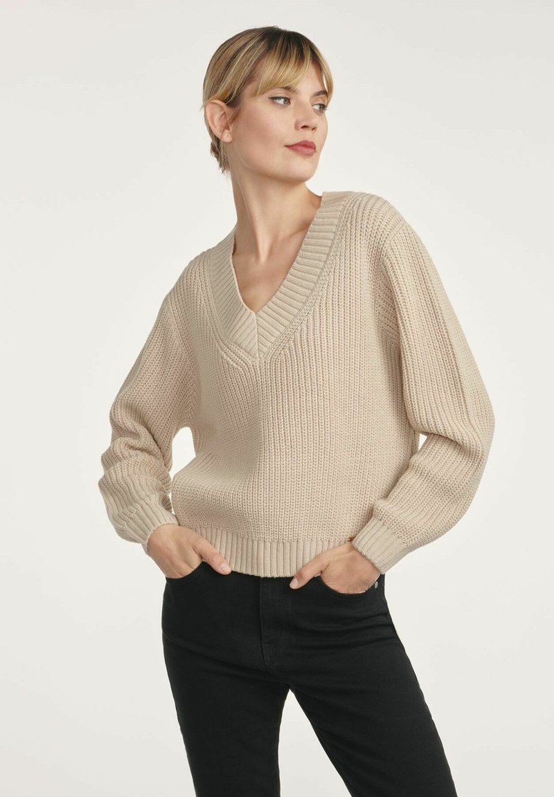 Kobieta w beżowym, dzianinowym swetrze z dekoltem w serek i czarnych spodniach, pozująca z rękami w kieszeniach, patrząca na bok na jednolitym tle.