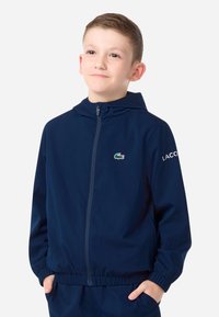 Tmavomodrá bunda na zips s kapucňou, s malým zeleným logom krokodíla na hrudi a nápisom „LACOSTE“ na rukáve. Elastické manžety.