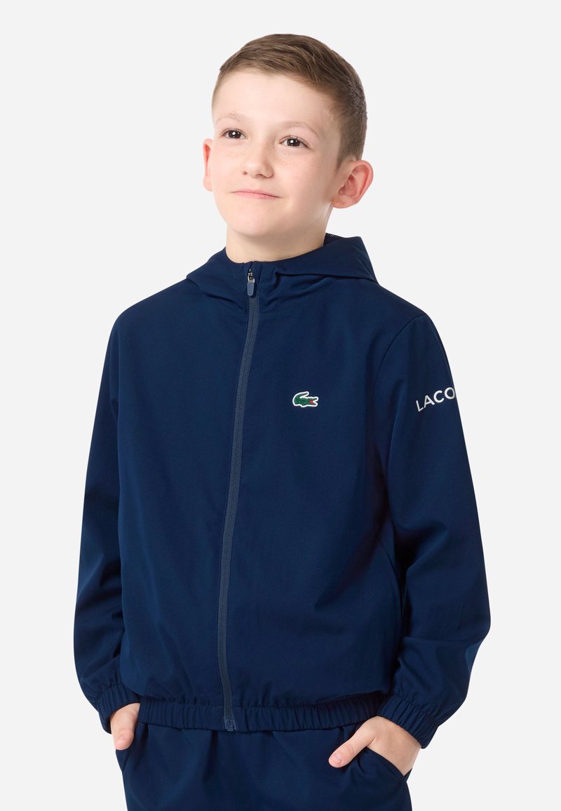 Tmavomodrá bunda na zips s kapucňou, s malým zeleným logom krokodíla na hrudi a nápisom „LACOSTE“ na rukáve. Elastické manžety.