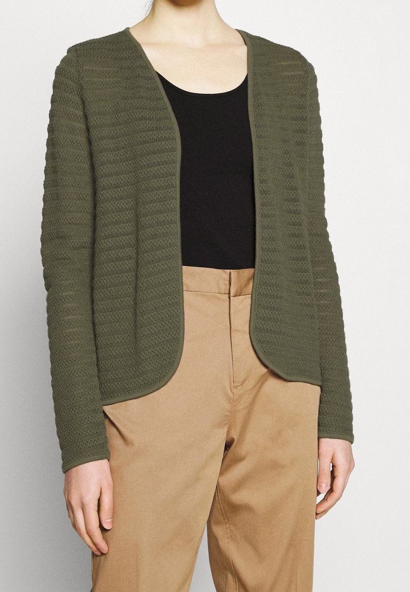 Femme portant un cardigan texturé vert olive ouvert, un haut noir et un pantalon beige taille haute sur un fond uni.