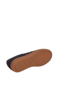Zapatilla negra con suela de goma marrón, mostrada desde la vista inferior destacando el patrón de textura de la suela y los detalles de costura.