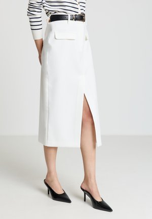 Jupe midi blanche en tissu lisse, avec une fente à l'avant, des poches latérales et une taille haute, accompagnée de mules noires à talons hauts et bouts pointus.