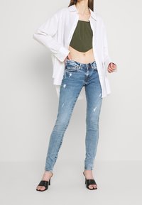 Camisa blanca de manga larga sobre un top corto de color verde oliva, combinada con jeans ajustados de mezclilla claros y desgastados, y mules negros de tacón.