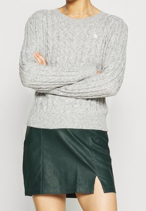 Femme les bras croisés portant un pull en maille torsadée gris clair et une mini-jupe en cuir vert foncé avec une fente à l'avant.