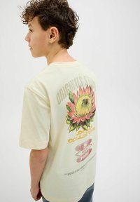 Gaiši dzeltena kokvilnas T-kreklu ar lielu saulespuķes grafiku uz aizmugures, un tekstu, kas ietver "ORIGINALS STUDIO" un "LOCAL COLLECTIVE."