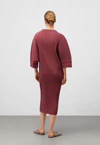 Rippkleid in Burgunderrot mit mittlerer Länge, breiten Ärmeln und lockerer Silhouette. Das Material hat eine strukturierte Oberfläche. Kombiniert mit beigen Slippern.