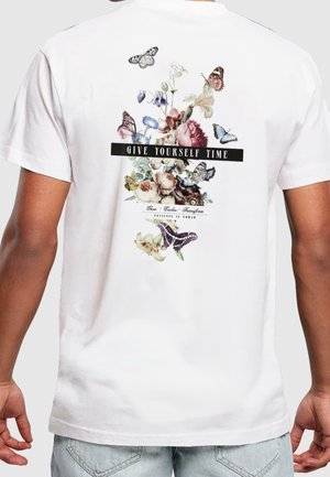 Hvid t-shirt med blomstret og sommerfugledesign på bagsiden og teksten "Give Yourself Time" med fede bogstaver.