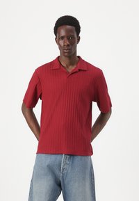 Rød ribbet polo-shirt med flad krave og korte ærmer, parret med lyseblå denimjeans. Tekstureret stof synligt over det hele.