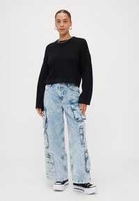 Zwarte gebreide trui met wijde mouwen gecombineerd met lichtblauwe denim cargobroek met meerdere zakken en een losvallende pasvorm. Zwarte sneakers.