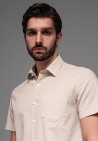 Lichtbeige kortärmige shirt van katoenmix, met een klassieke kraag, borstzak en witte knopen tegen een neutrale achtergrond.