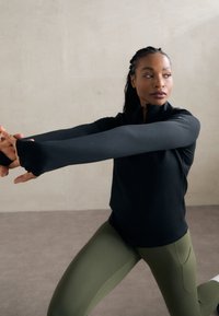 Top sportiv negru cu fermoar, cu mâneci lungi și găuri pentru degete, combinat cu colanți strâmți de culoare verde măslinie, într-un decor interior cu nuanțe neutre.