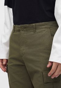 Primo piano di una persona che indossa pantaloni cargo verde oliva con bottone e tasca frontale, abbinati a una maglietta nera e maniche lunghe bianche visibili.