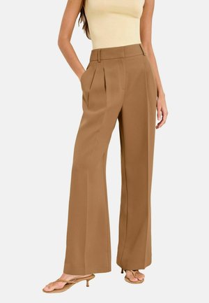Femme portant un pantalon marron à taille haute, large aux jambes et plissé, associé à un haut beige sans manches et des sandales marron à talons.
