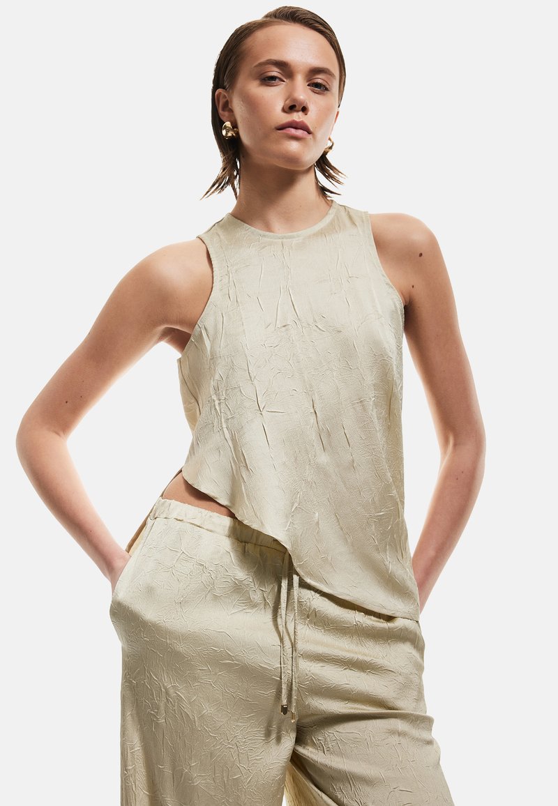 Top senza maniche color beige chiaro con finitura texturizzata e orlo asimmetrico, abbinato a pantaloni texturizzati coordinati con vita elastica.