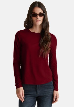 Femme avec de longs cheveux châtains ondulés portant des lunettes de soleil écaille de tortue, un haut à manches longues bordeaux et un jean bleu foncé, se tenant devant un fond blanc.
