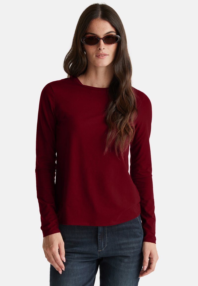 Frau mit langen, welligen braunen Haaren, die Schildkrötensonnenbrille, ein bordeauxfarbenes Langarmshirt und dunkelblaue Jeans trägt, steht vor einem weißen Hintergrund.