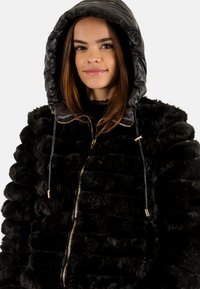 Zwarte faux fur ritsjack met een gevoerde capuchon, voorzien van een gestructureerd patroon, gouden hardware en trekkoorden voor een verstelbare pasvorm.