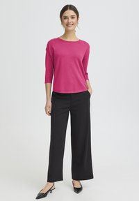 Pull en maille fuchsia à manches trois quarts, avec des boutons sur l'épaule, associé à un pantalon noir à jambes larges et des chaussures à talons noires.