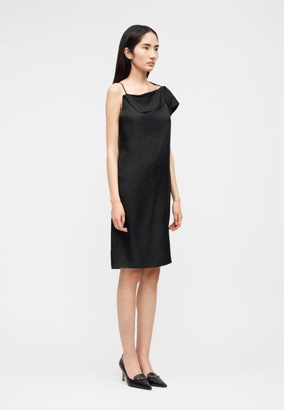 Modèle féminin debout, portant une robe en satin noire mi-longue avec des fines bretelles et un décolleté asymétrique, associée à des talons pointus noirs.