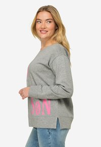 Grå sweatshirt med rosa text framtill, med en avslappnad passform, bred halsringning och sidospår. Materialet verkar mjukt och strukturärt.