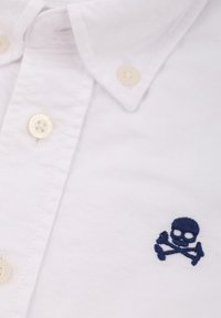 Weißes Hemd mit Knopfverschluss, das beige Knöpfe und ein dunkelblaues gesticktes Totenkopf- und Kreuzboneslogo auf der linken Brustseite aufweist.