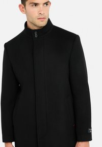 Cappotto nero in lana con collo alto, dotato di due bottoni frontali e tasche laterali. Tessuto liscio e vestibilità sartoriale, con dettaglio logo sulla manica.