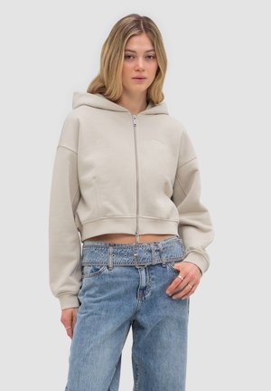 Junge Frau trägt einen kurzen beigen Kapuzenpullover mit Reißverschluss und hoch taillierte blaue Jeans mit breitem Jeansgürtel, steht vor einem einfarbigen Hintergrund.