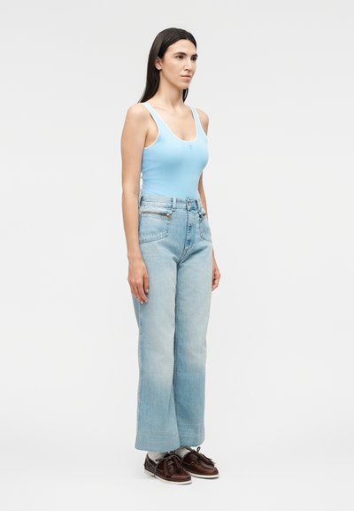 Γυναίκα όρθια, φορώντας ανοιχτό μπλε αμάνικη μπλούζα, ψηλόμεσα φαρδιά ανοιχτόχρωμα τζιν, λευκές κάλτσες και σκουρόκαφέ μοκασίνια με φούντες.